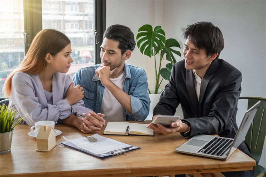 Three people sit around a table with documents, a notebook, a laptop, and a calculator, having a discussion in a well-lit office setting.