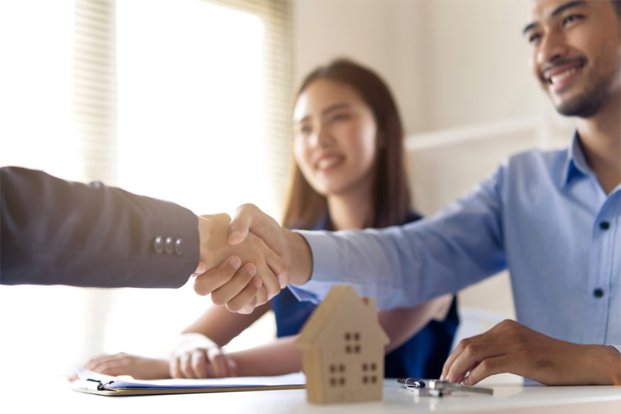 Two people shake hands across a table with a smiling woman watching and a small model house and keys in the foreground.