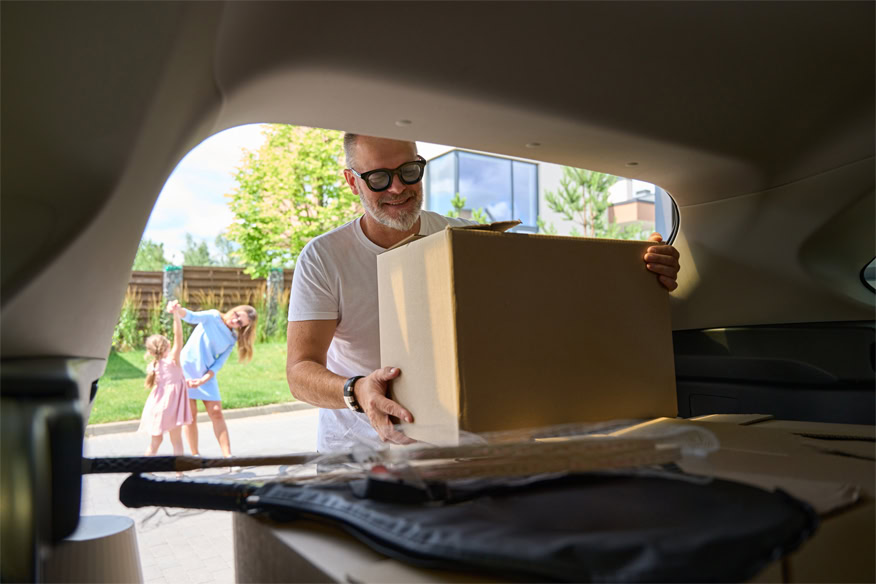 A man loads a cardboard box into a car trunk while a woman and two children stand outside in the background.