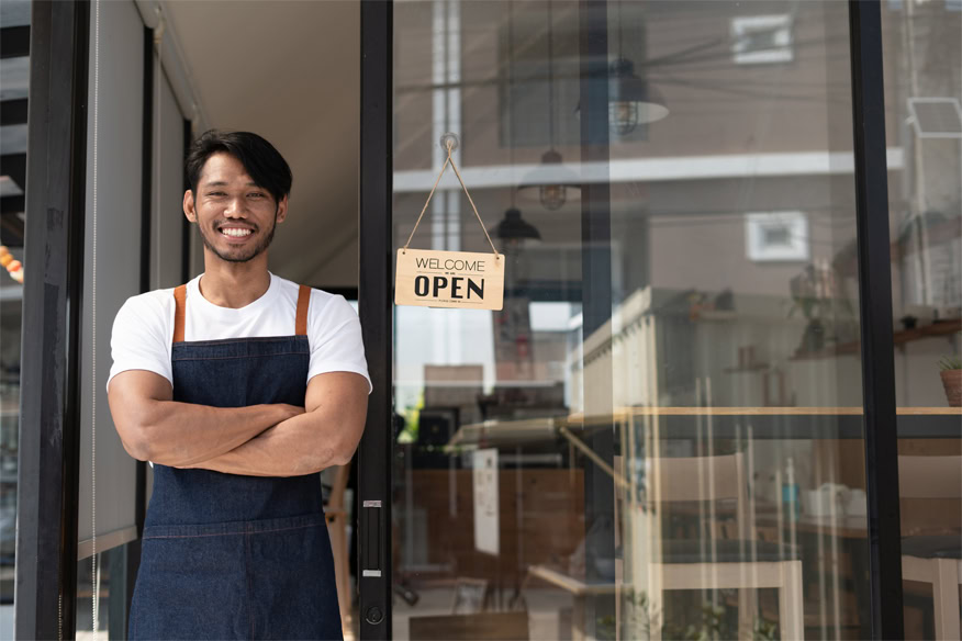 A man wearing an apron stands with folded arms in front of a glass door with a "Welcome, Open" sign, likely at the entrance of a store or café.