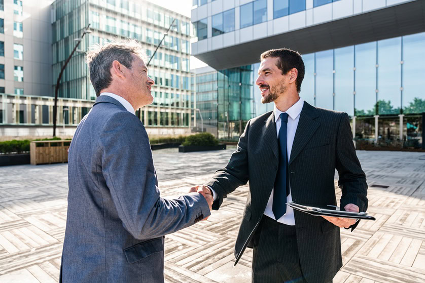 Two men in business suits shake hands outside a modern office building, as one holds a tablet and mortgage pre-approval documents.