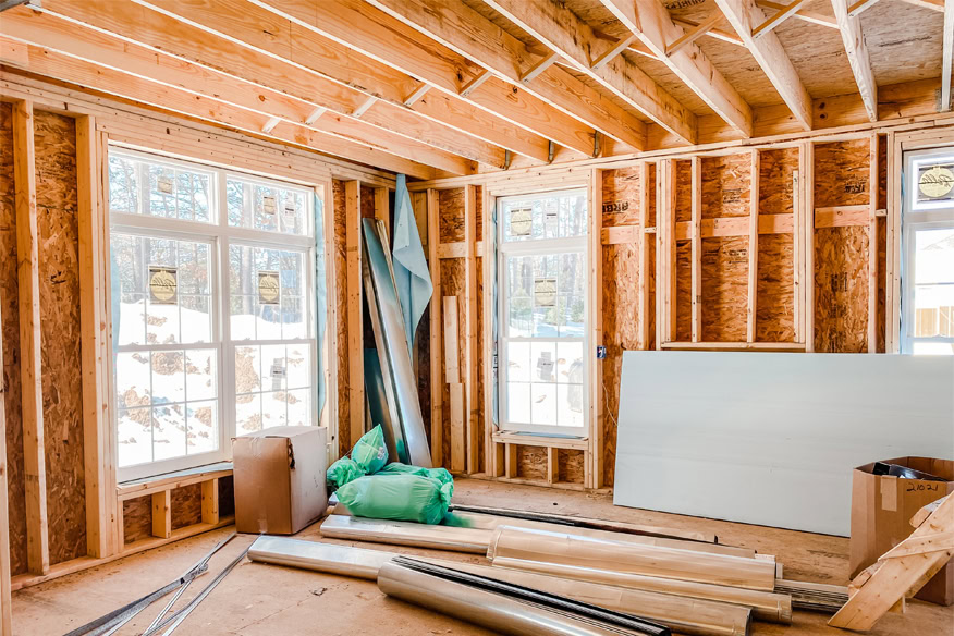 Unfinished room with exposed wooden framing, two large windows, and construction materials scattered on the floor.
