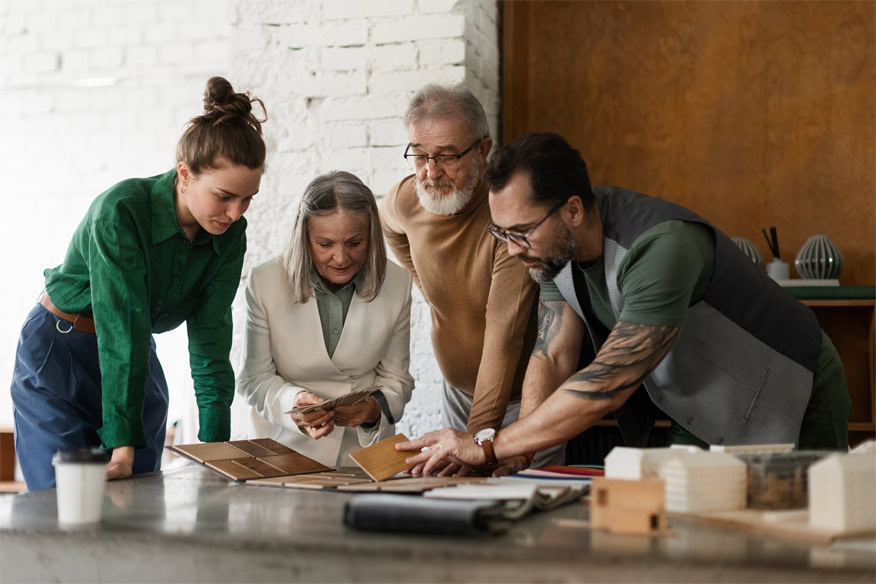 Four people stand around a table examining material samples and architectural models in a modern workspace.