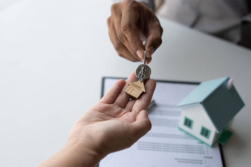 A person hands over a key with a house-shaped keychain to another person, with a document and a small model house on the table.