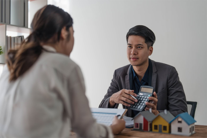 A man in a suit holds a calculator while talking to a woman across a desk with small model houses on it.
