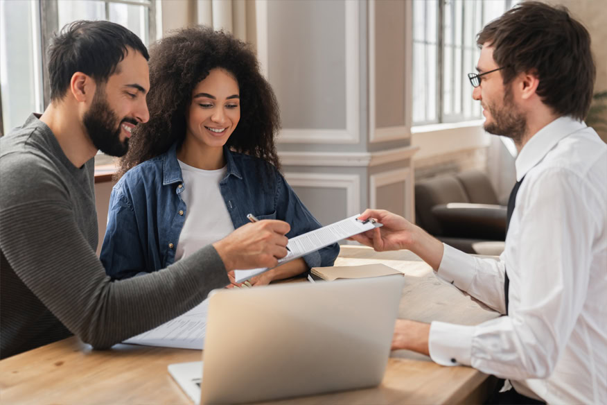 Three people sit at a table with a laptop and documents. One person hands a paper to another, while the third person smiles and observes.