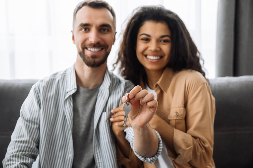 A smiling couple sits on a couch, with one person holding a house key toward the camera, suggesting they have acquired a new home.