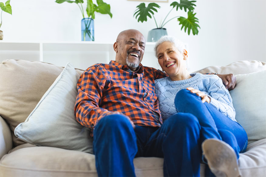 An elderly couple sits close together on a beige couch, smiling and relaxed, with green houseplants in the background.
