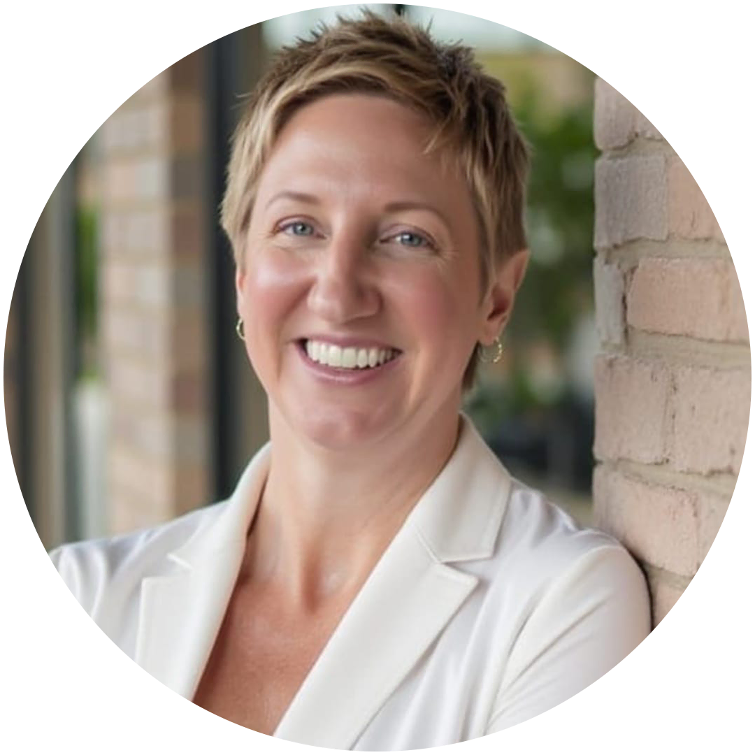 Smiling person with short blonde hair wearing a white blazer, standing indoors and leaning against a brick wall, ready to offer expert mortgage solutions.