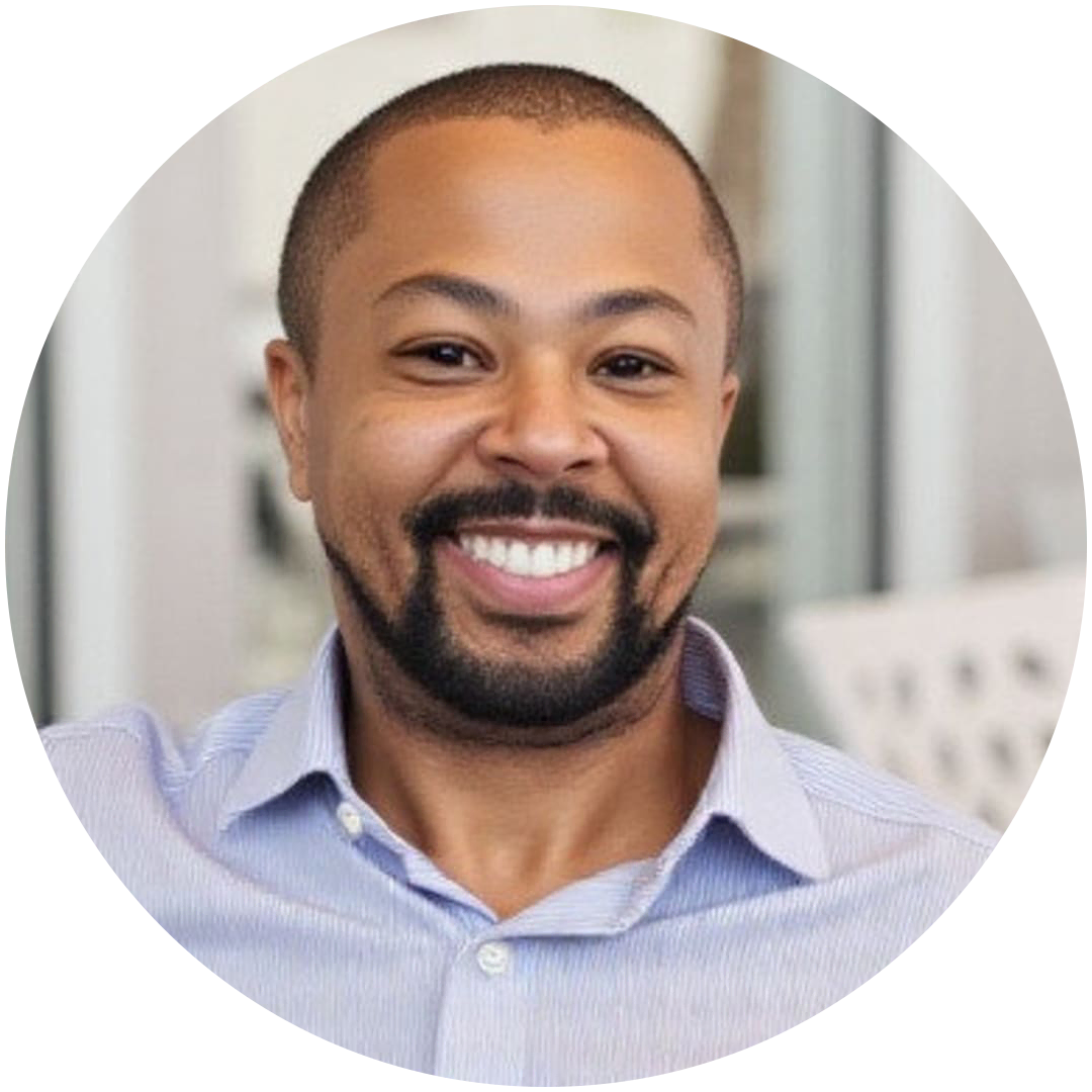 A man with a short beard and mustache smiles at the camera, wearing a light blue collared shirt in an indoor office setting, ready to discuss mortgage solutions.