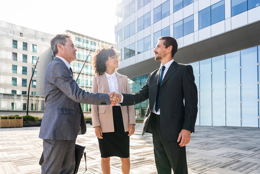 Two men in business attire shake hands outdoors in front of a modern office building, finalizing a mortgage-financing deal, while a woman stands beside them, smiling.
