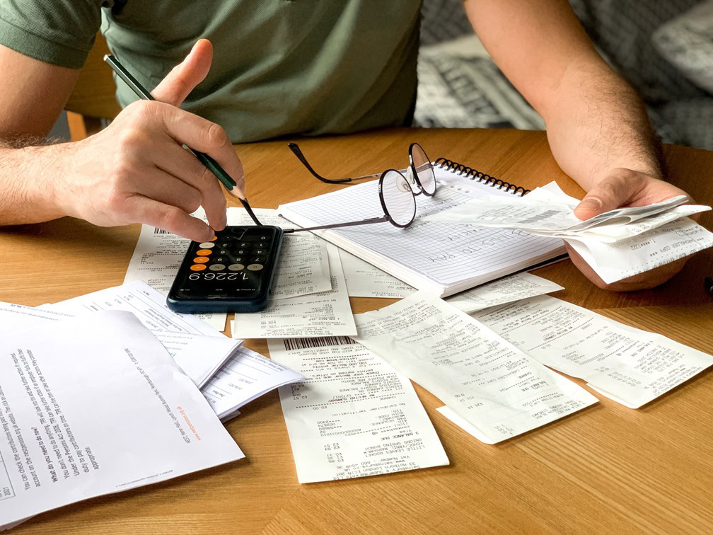 A person uses a calculator and pencil at a table covered with receipts, bills, and a notebook—organizing expenses and reviewing mortgage-financing options, with eyeglasses resting on the paperwork.