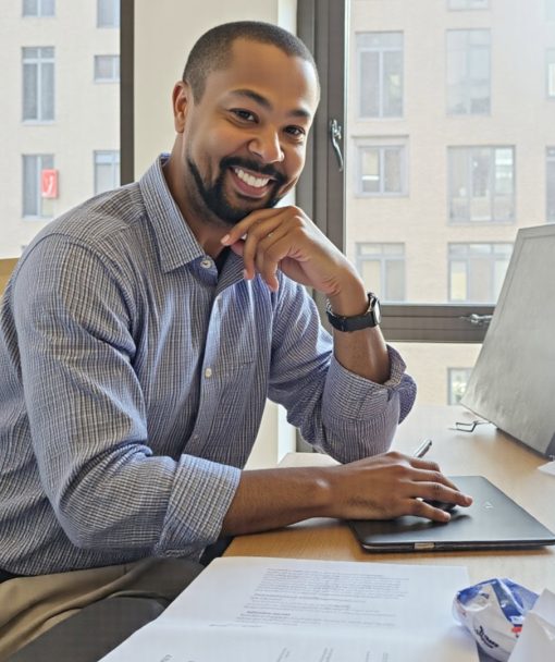 A man sits at a desk in an office, smiling at the camera, with a laptop, papers, and office supplies in front of him as he discusses mortgage solutions.